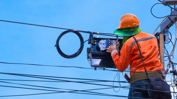 Rear view of technician on wooden ladder checking code numbers of fiber optic cable lines in internet splitter box for repairing to work normally on electric pole against a blue sky background